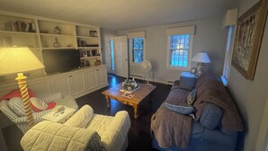 Una sala de estar con un sofá, sillones, una mesa de centro y un televisor. Hay estanterías con libros y objetos decorativos, y una puerta que da a otra habitación.