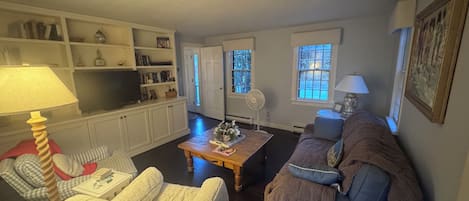 Un salon comprenant un canapé, des fauteuils, une table basse et un téléviseur. On y trouve également des étagères remplies de livres et d’objets décoratifs, ainsi qu’une porte donnant sur une autre pièce.