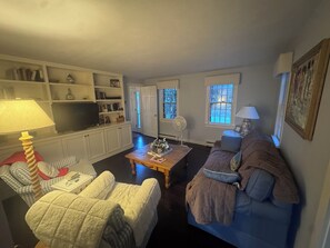 Ein Wohnzimmer mit einer Couch, Sesseln, einem Couchtisch und einem Fernseher. Regale mit Büchern und Dekoration, und eine Tür zu einem anderen Raum.