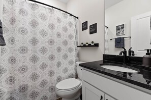 Un baño con cortina de ducha estampada, inodoro blanco, lavabo negro con espejo y estante con plantas.