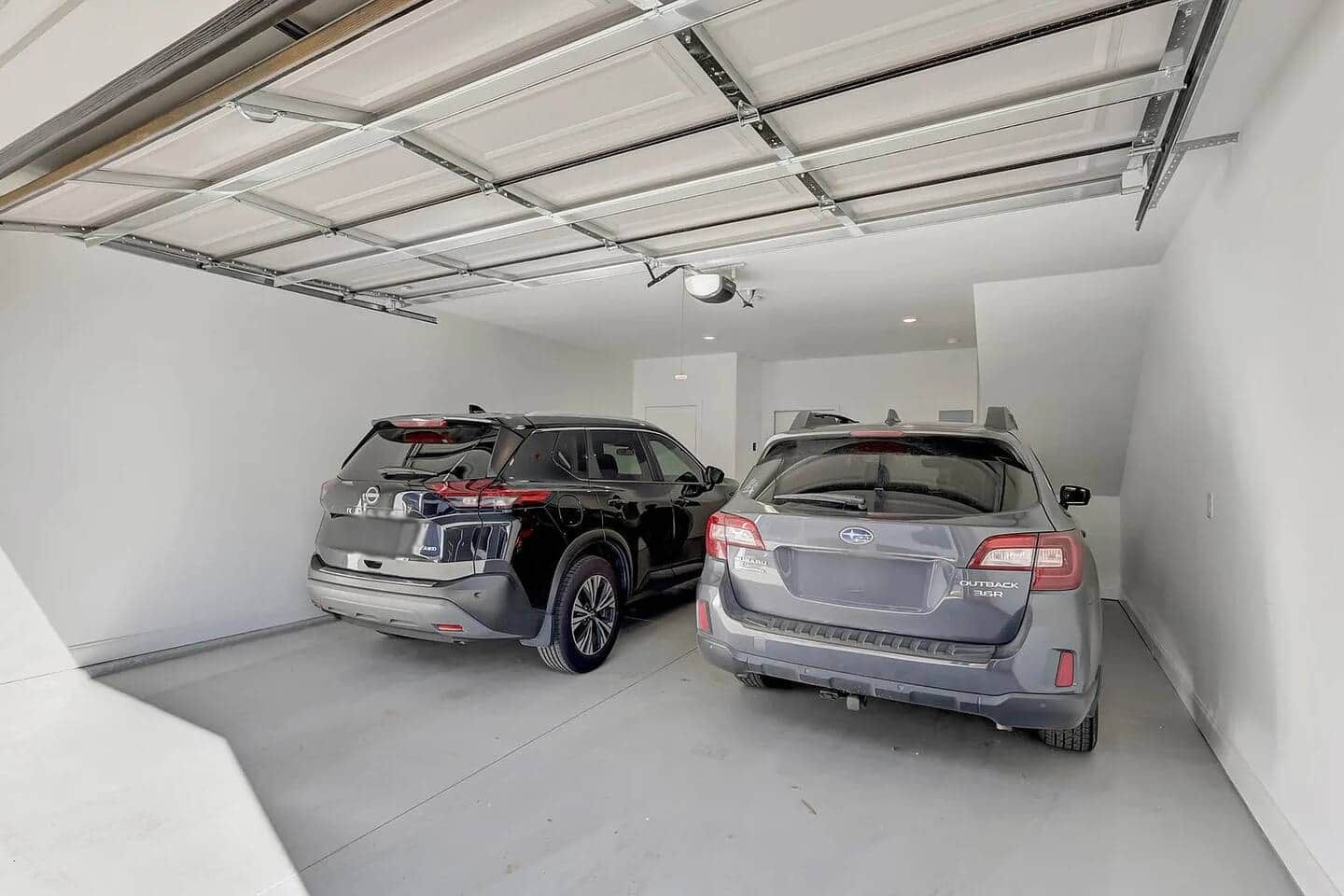 A garage with two cars parked, a white wall, and a ceiling with metal beams.