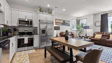 A modern kitchen with stainless steel appliances, a dining area with a wooden table and bench, a living room with a brown leather sofa, and a large window with curtains.