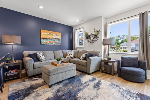 A modern living room with a sectional sofa, a coffee table, and a side table with a lamp. There is a large window with curtains, a wall-mounted plant, and a framed picture.