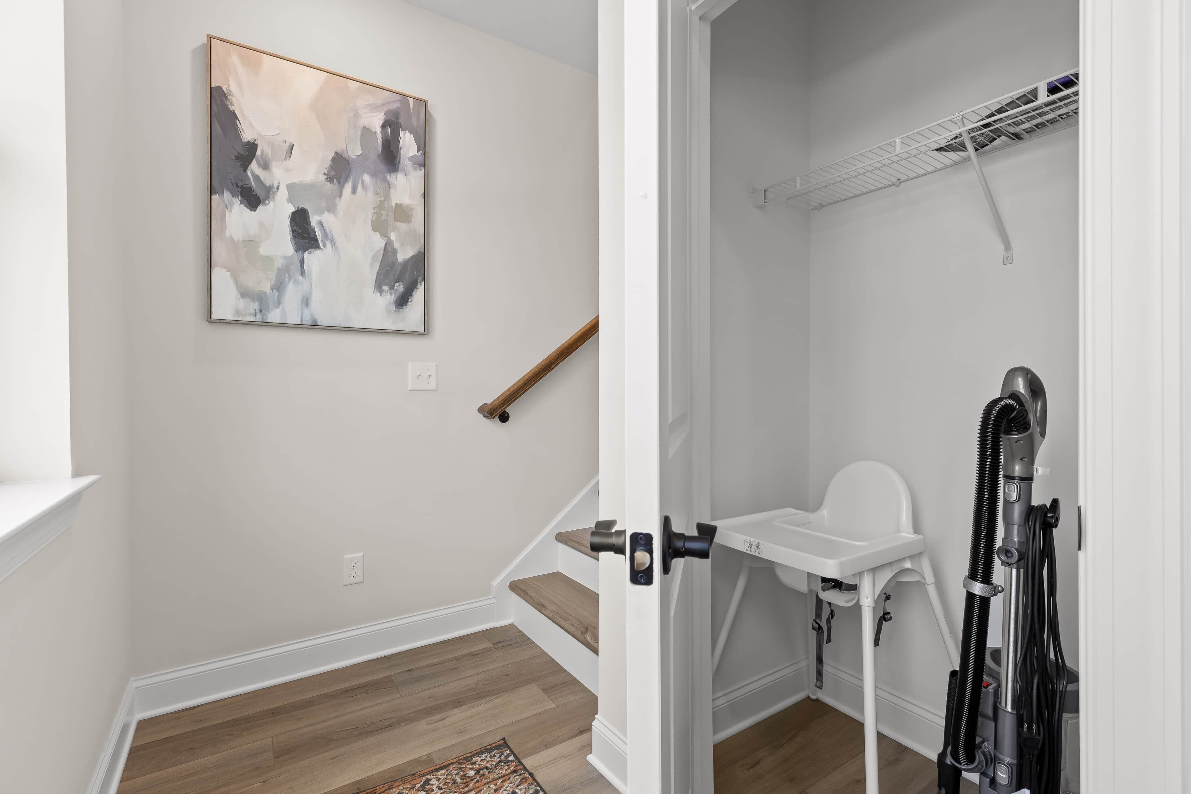 A room with a wooden floor, a white wall, a painting, a wooden stick leaning against the wall, and a white chair with a vacuum cleaner.