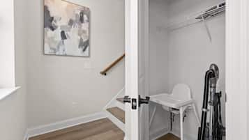 A room with a wooden floor, a white wall, a painting, a wooden stick leaning against the wall, and a white chair with a vacuum cleaner.