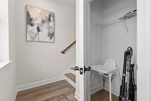 A room with a wooden floor, a white wall, a painting, a wooden stick leaning against the wall, and a white chair with a vacuum cleaner. - ChattVistas|Dwntwn Townhouse|Deck Fire Pit|2Bdrm (Chattanooga)
