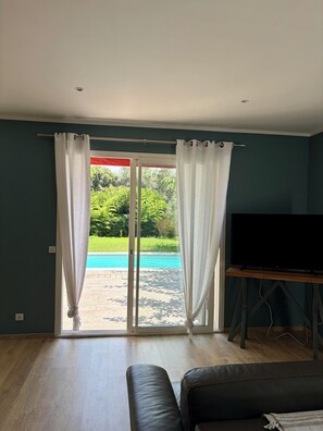 A room with a television on a wooden stand, a black leather sofa, and a view of a swimming pool through a glass door with white curtains. - Villa in Saint-Florent with a pool, 5 minutes from the beach (by car) (Oletta)