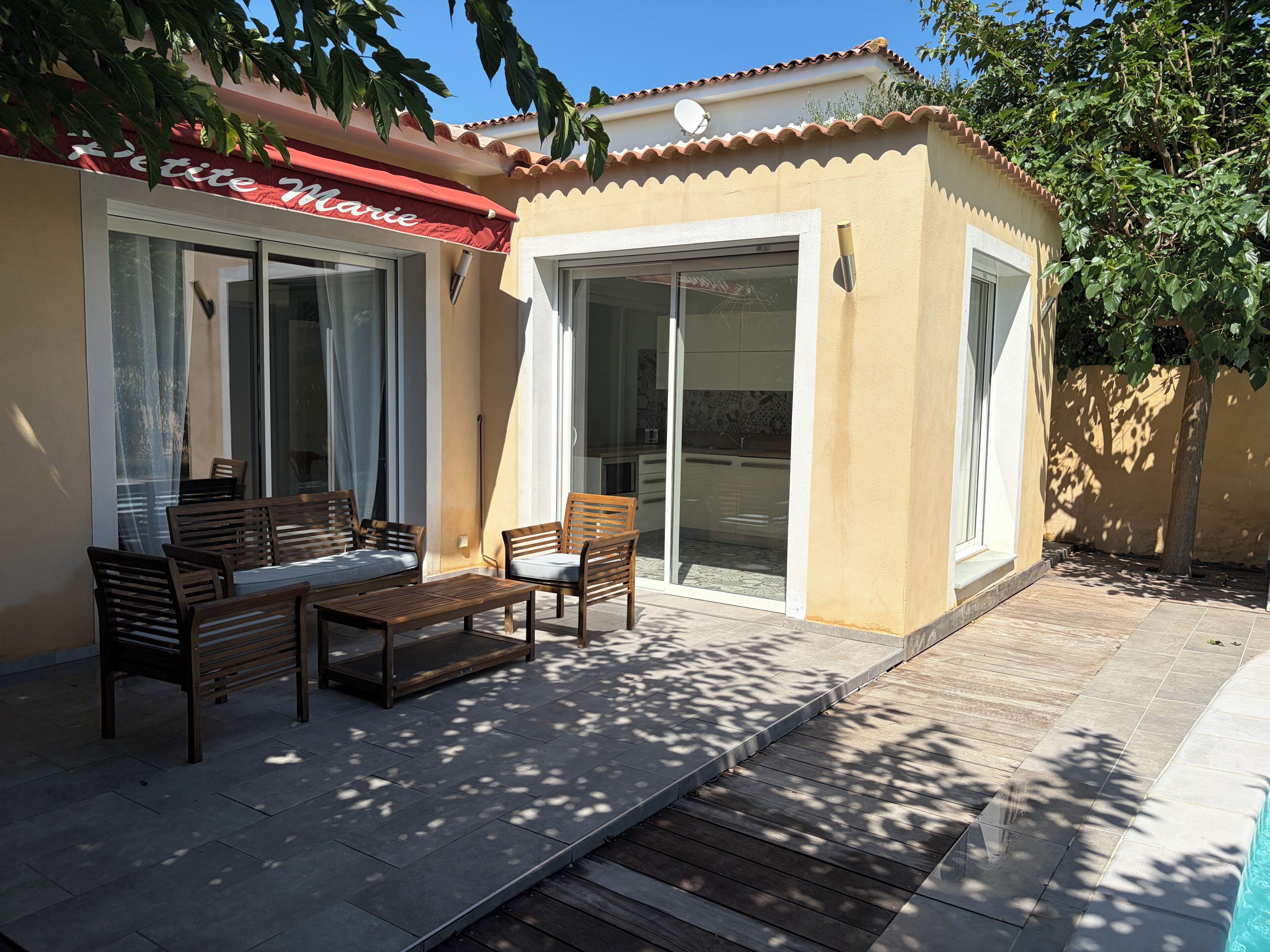 Un espace patio aménagé avec du mobilier d’extérieur, une porte-fenêtre coulissante donnant sur une cuisine et un bâtiment doté d’un auvent rouge.