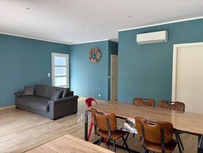 A modern living space with a grey sofa, a wooden dining table with chairs, a clock on the wall, and an air conditioning unit. - Villa in Saint-Florent with a pool, 5 minutes from the beach (by car) (Oletta)