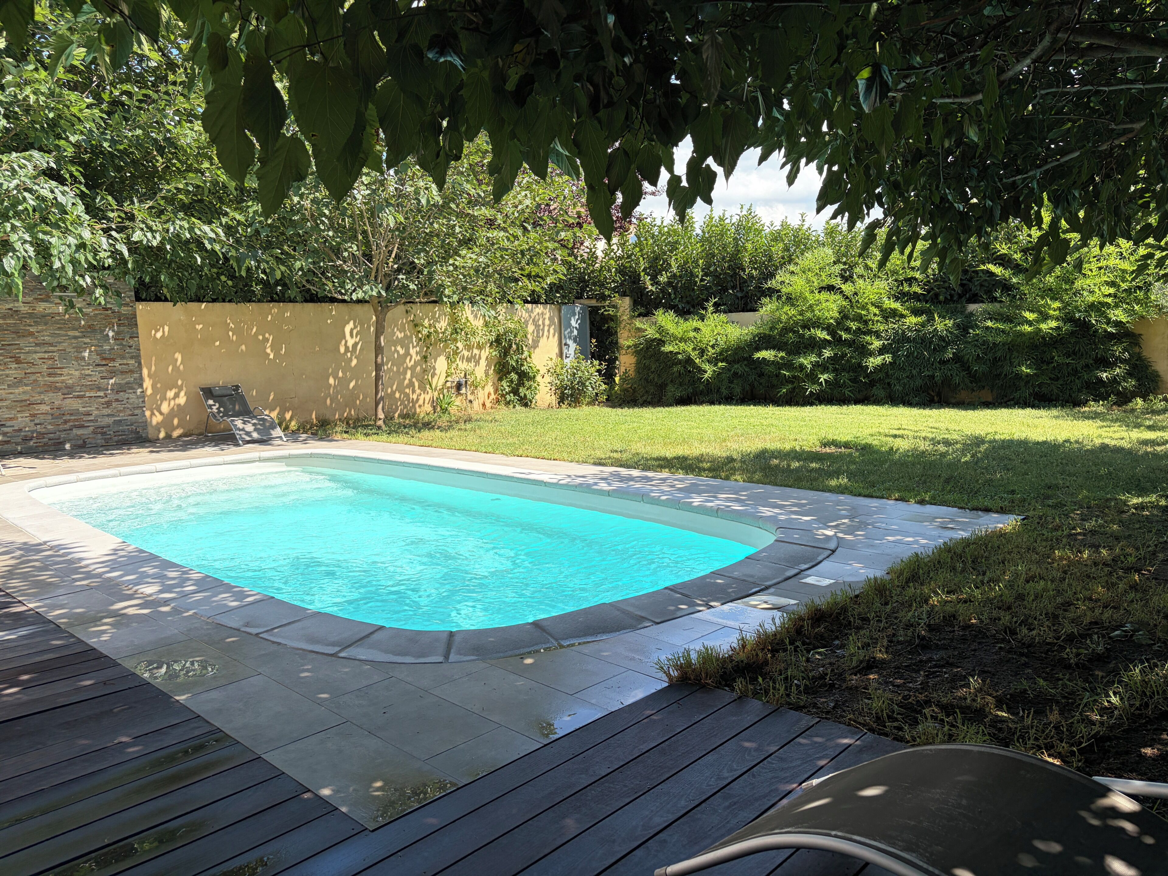 A rectangular swimming pool with a wooden deck surrounding it, set in a backyard with a stone wall and lush greenery.