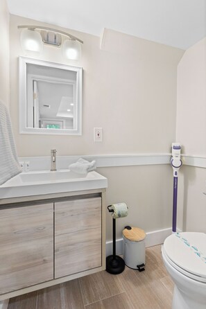 Een badkamer met een wit wastafelmeubel, spiegel en toilet. Er staat een prullenbak en een dweil.