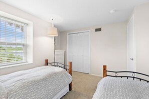 Ein Schlafzimmer mit zwei Betten, einem Fenster mit Jalousien, einer Nachttischlampe, einem Kleiderschrank und einer Tür.