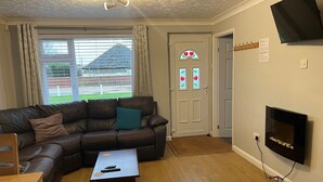 A living room with a leather sofa, a coffee table, a flat-screen TV mounted on the wall, and a door with a stained glass window. - These quality bungalows are situated 200mt from Hemsby beaches and all ammenties (Gt Yarmouth)