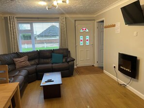 A living room with a leather sofa, a coffee table, a flat-screen TV mounted on the wall, and a door with a stained glass window. - These quality bungalows are situated 200mt from Hemsby beaches and all ammenties (Gt Yarmouth)