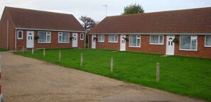 A row of brick houses with white doors and windows, a gravel driveway, and a grassy lawn. - These quality bungalows are situated 200mt from Hemsby beaches and all ammenties (Gt Yarmouth)