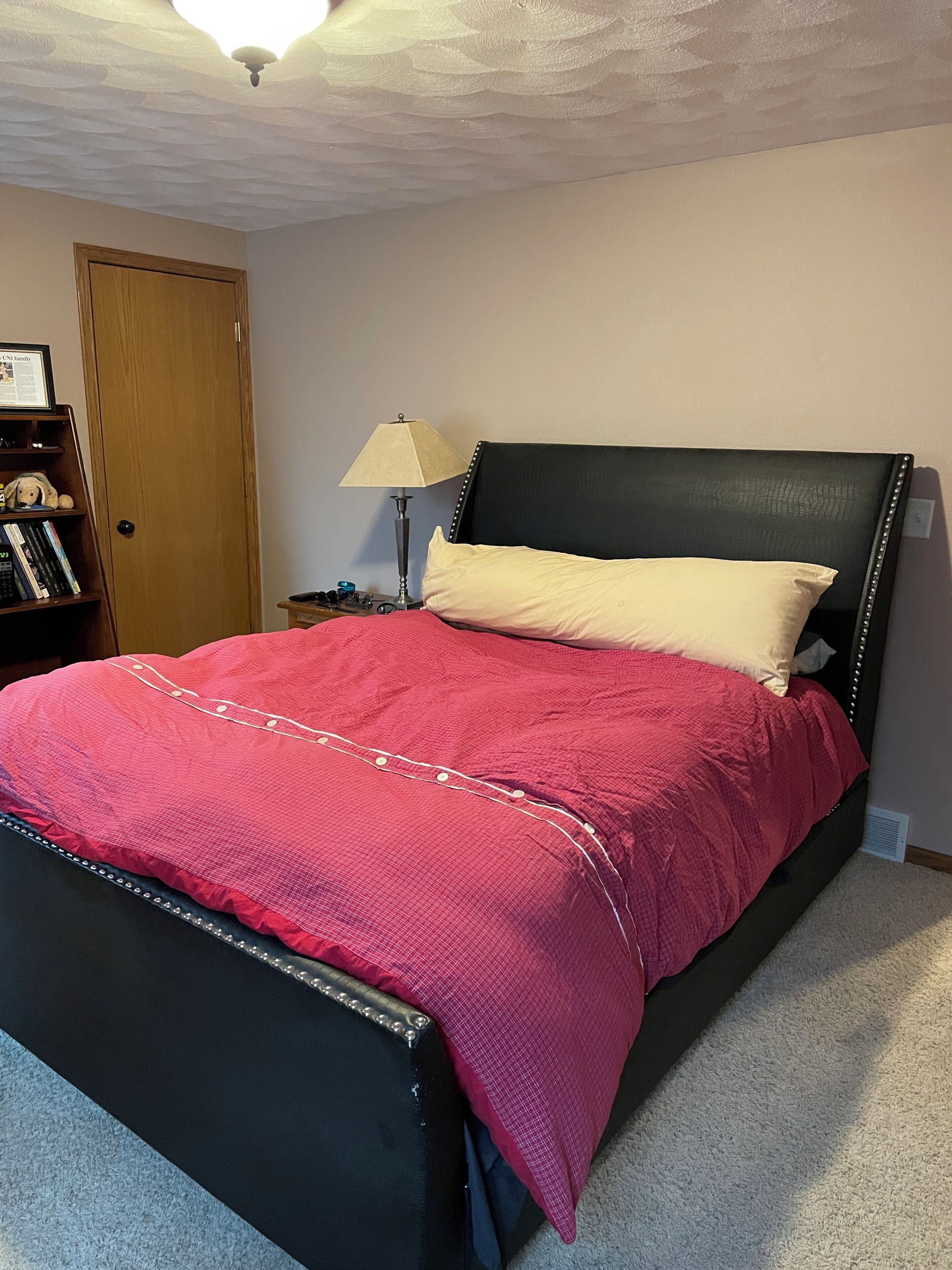 A bedroom with a bed, a nightstand, a lamp, a bookshelf, and a door.