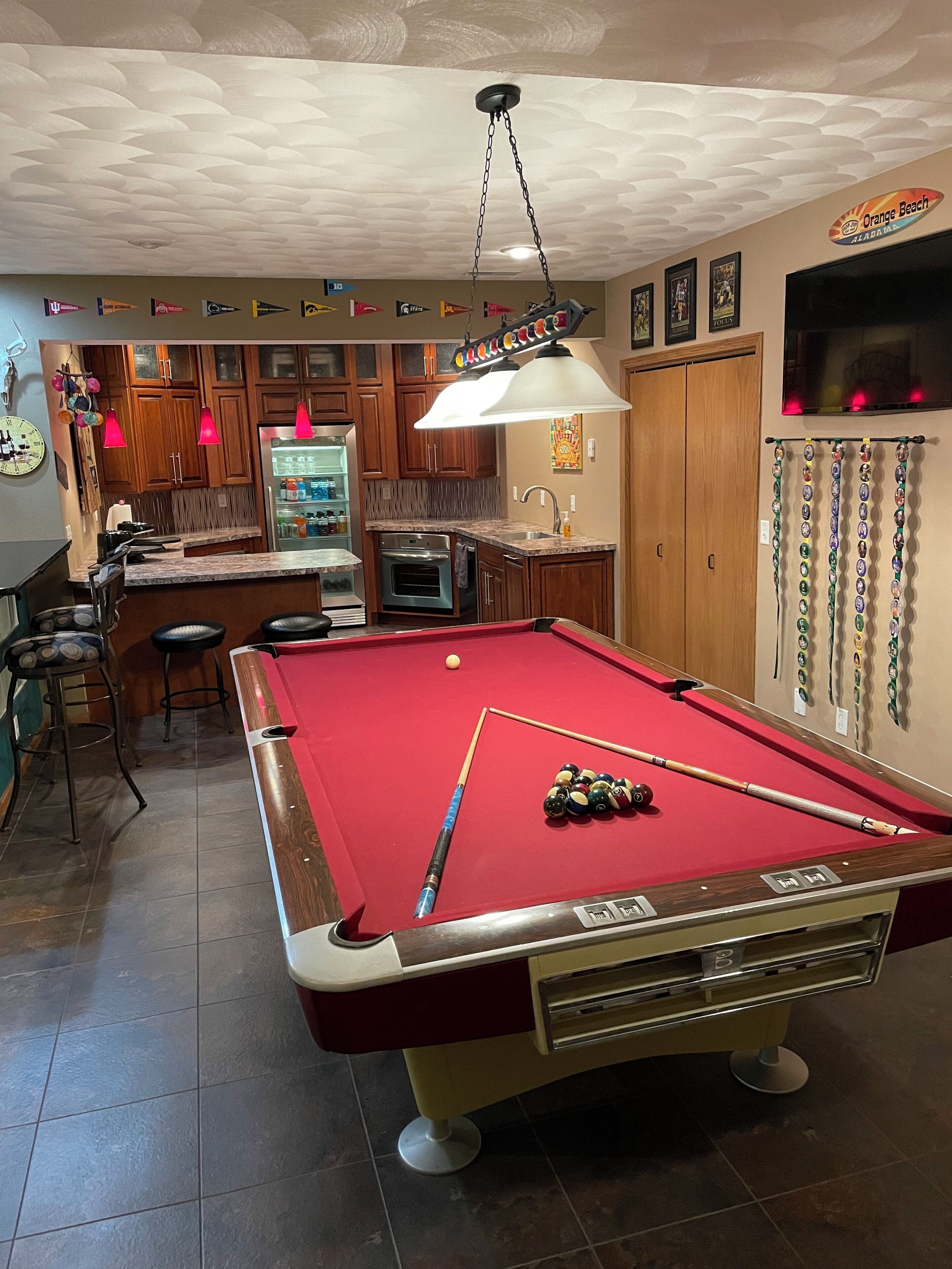 A pool table with a red felt surface, a bar area with stools, and a kitchen with wooden cabinets.