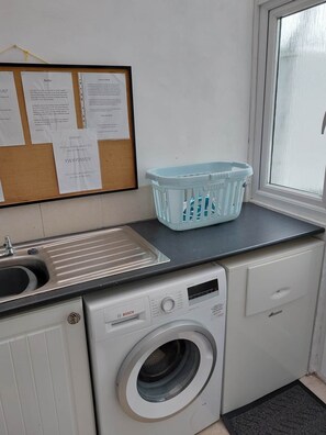 Ein Waschraum mit Waschmaschine, Waschbecken, Schwarzes Brett mit Aushängen und einem Fenster.