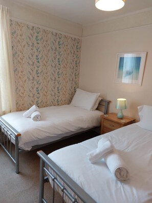A hotel room with two single beds, a nightstand with a lamp, a framed picture on the wall, and a window with curtains. - 14 Channel View, St. Ives, Cornwall TR26 1RG (St Ives)