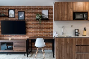 A modern kitchen with a brick wall, wooden cabinets, a flat-screen TV, a dining area with a white chair, and a coffee machine. - ENZO Privé | Studios - Modern Executive Suite in Laureles (Medellín)