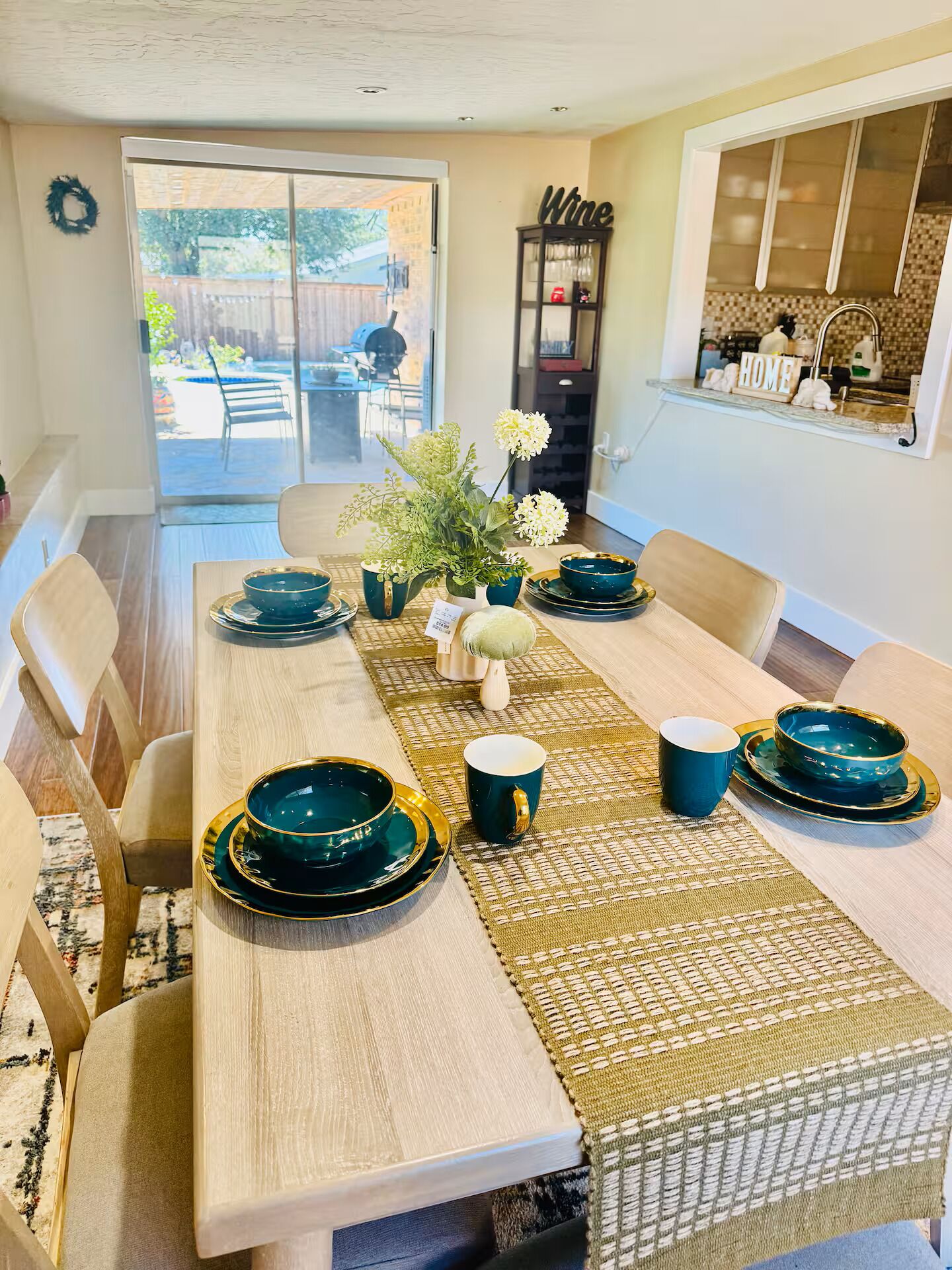 A dining area with a long table set for six, featuring teal and gold dinnerware, a woven placemat, and a vase with flowers.