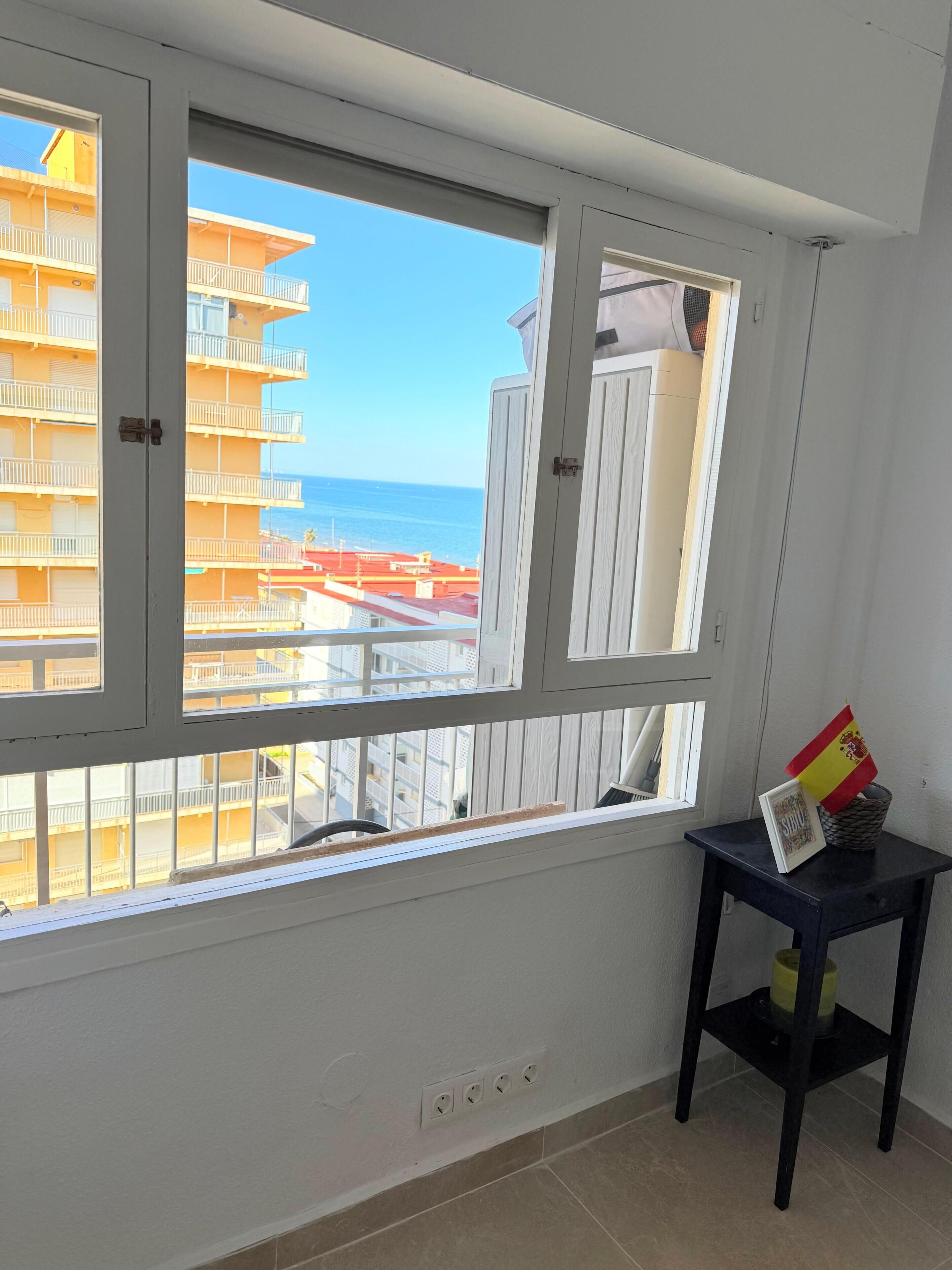 A room with a large window overlooking a building with balconies and a view of the sea. There is a small table with a flag and a picture frame on it.