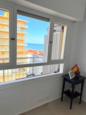 Une pièce dotée d’une grande fenêtre donnant sur un immeuble avec des balcons et offrant une vue sur la mer. On y trouve une petite table sur laquelle sont posés un drapeau et un cadre photo.