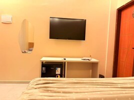 A hotel room with a bed, a flat-screen TV mounted on the wall, a mirror, a small desk with a computer, and a door.