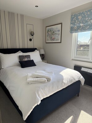 A neatly made bed with pillows and a folded towel, a bedside lamp, a framed picture on the wall, and a window with a view of buildings. -  10mins  drive from Terminal 5 & 35 mins train to London Waterloo (Staines-upon-Thames)