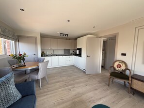 A modern kitchen with white cabinets, a dining area with a wooden table and grey chairs, and a small seating area with a blue sofa and patterned cushions. -  10mins  drive from Terminal 5 & 35 mins train to London Waterloo (Staines-upon-Thames)