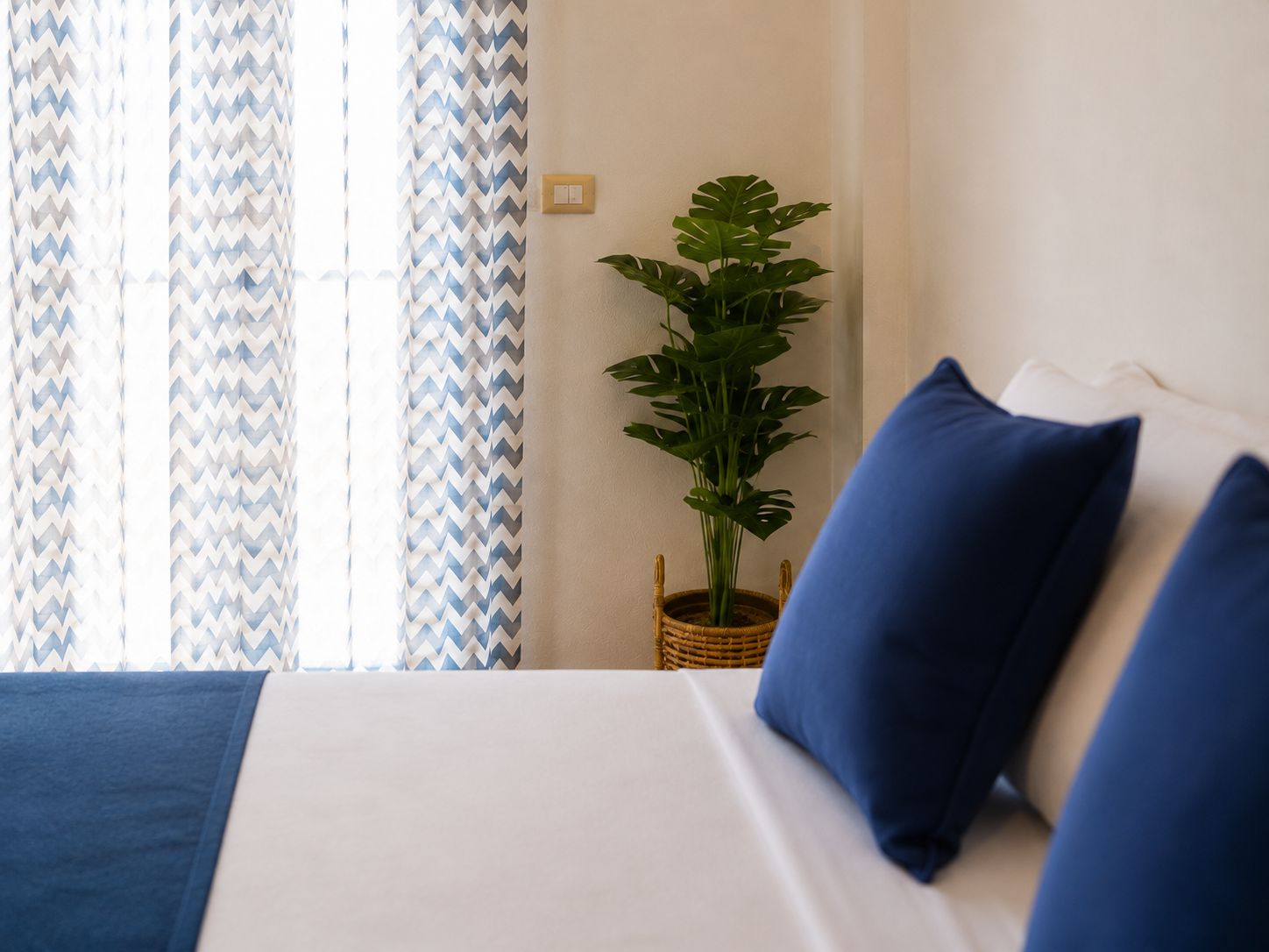 A bed with blue and white bedding, a potted plant, and a window with patterned curtains.