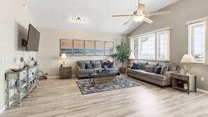 Un salon spacieux avec un grand canapé, une table basse et un ventilateur de plafond. Un téléviseur est fixé au mur et une fenêtre laisse entrer la lumière naturelle.