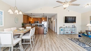 A spacious kitchen with wooden cabinets, a dining area with a table and chairs, a ceiling fan, and a television mounted on the wall. - Top-Floor Coastal Condo with Pool, Private Deck & Walk to the Beach (Wildwood Crest)