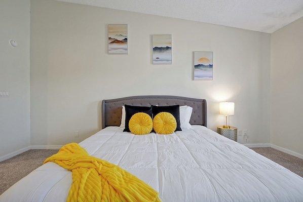 A neatly made bed with a white comforter and two decorative pillows, a bedside lamp, and three framed artworks on the wall.