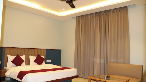 Una habitación de hotel con cama, mesita de noche, una silla, una mesa, ventilador de techo y un ventanal con cortinas.