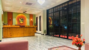 Zona de recepción de un hotel con un mostrador de madera, un cartel de recepción, una araña y un amplio ventanal, además de una alfombra roja.