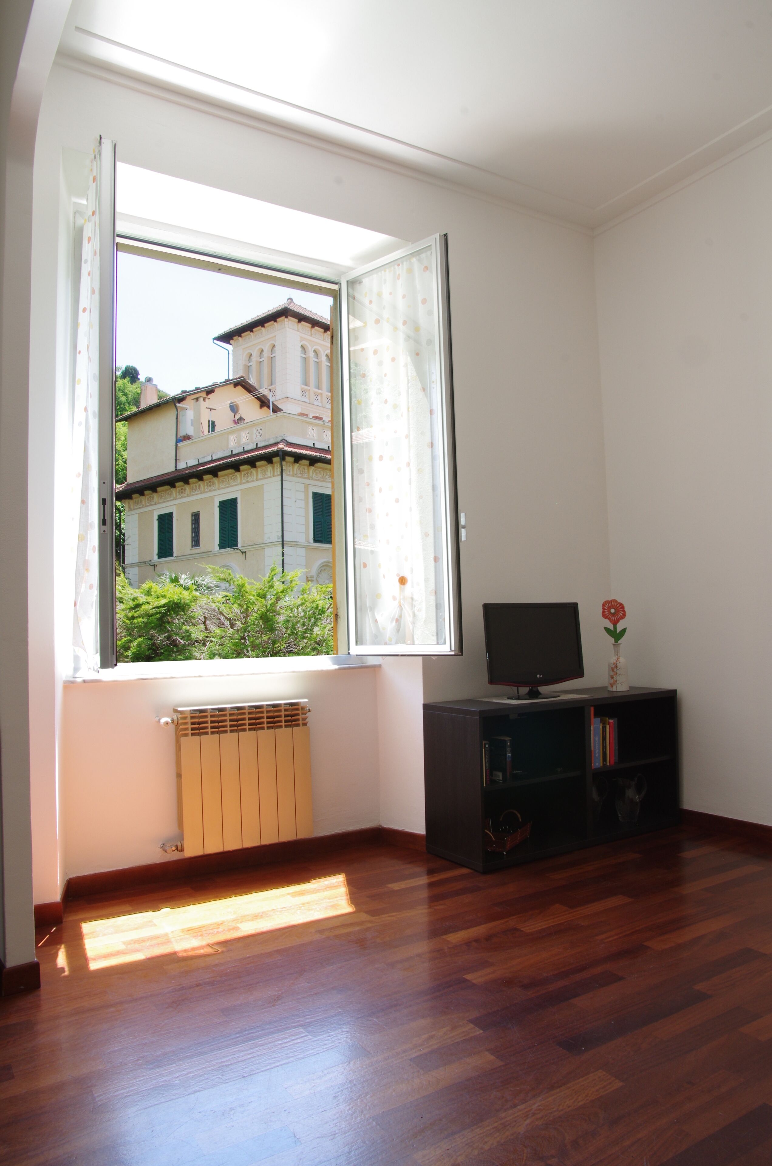 Una stanza con un'ampia finestra, un termosifone, una televisione su un mobile e una libreria.