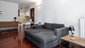 A modern living room with a grey sectional sofa, a wooden coffee table, and a dining area with white chairs. - Italianway Comfort - Vico Magistretti 1 B (Milano)