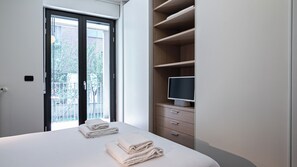 A hotel room with a bed, a TV on a wooden cabinet, and a balcony with a view of greenery. - Italianway Comfort - Vico Magistretti 1 B (Milano)