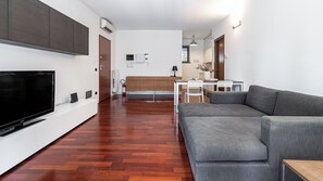 Ein modernes Wohnzimmer mit Fernseher, grauem Sofa, Holzboden, einem Essbereich mit Tisch und Stühlen sowie einer Küche im Hintergrund.