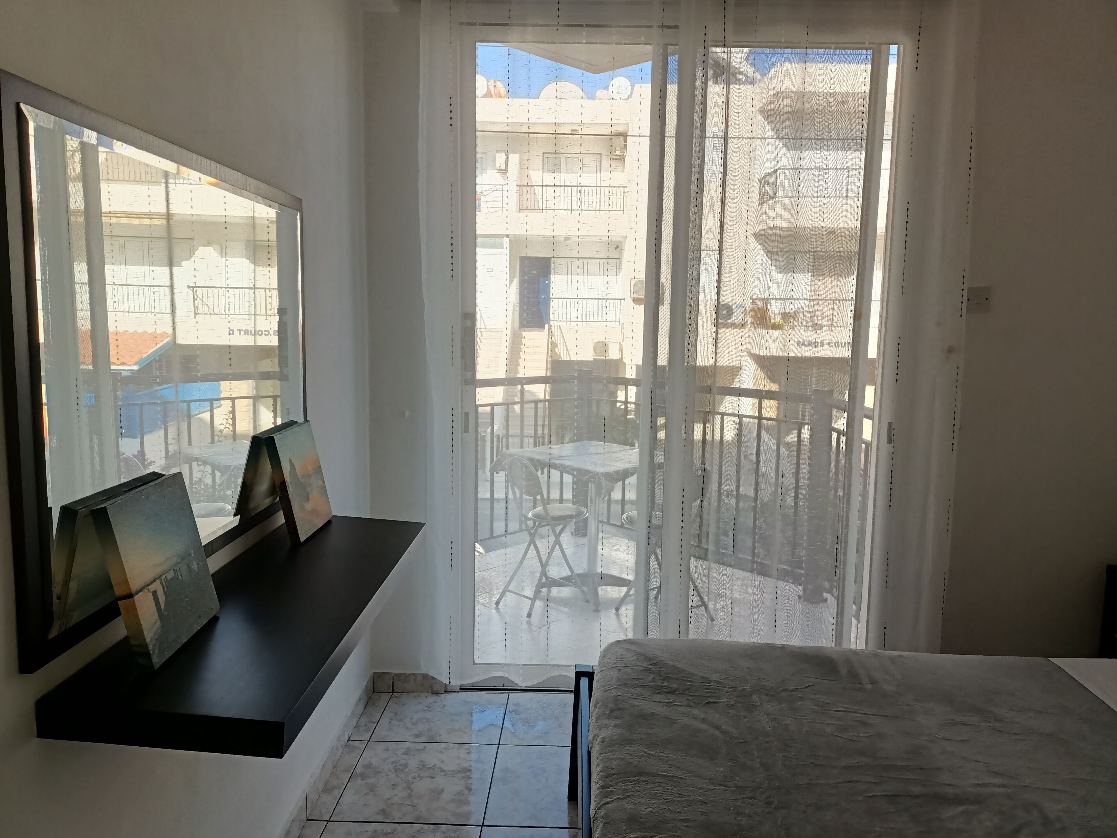 A room with a bed, a window with a view of the outside balcony, and a small table with a picture frame.