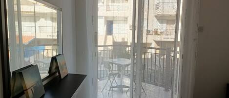 A room with a bed, a window with a view of the outside balcony, and a small table with a picture frame.