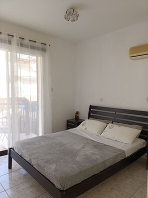 A bedroom with a bed, a nightstand, a wall-mounted air conditioner, and a view through a window with sheer curtains. - MOLOS Complex  (Pafos)