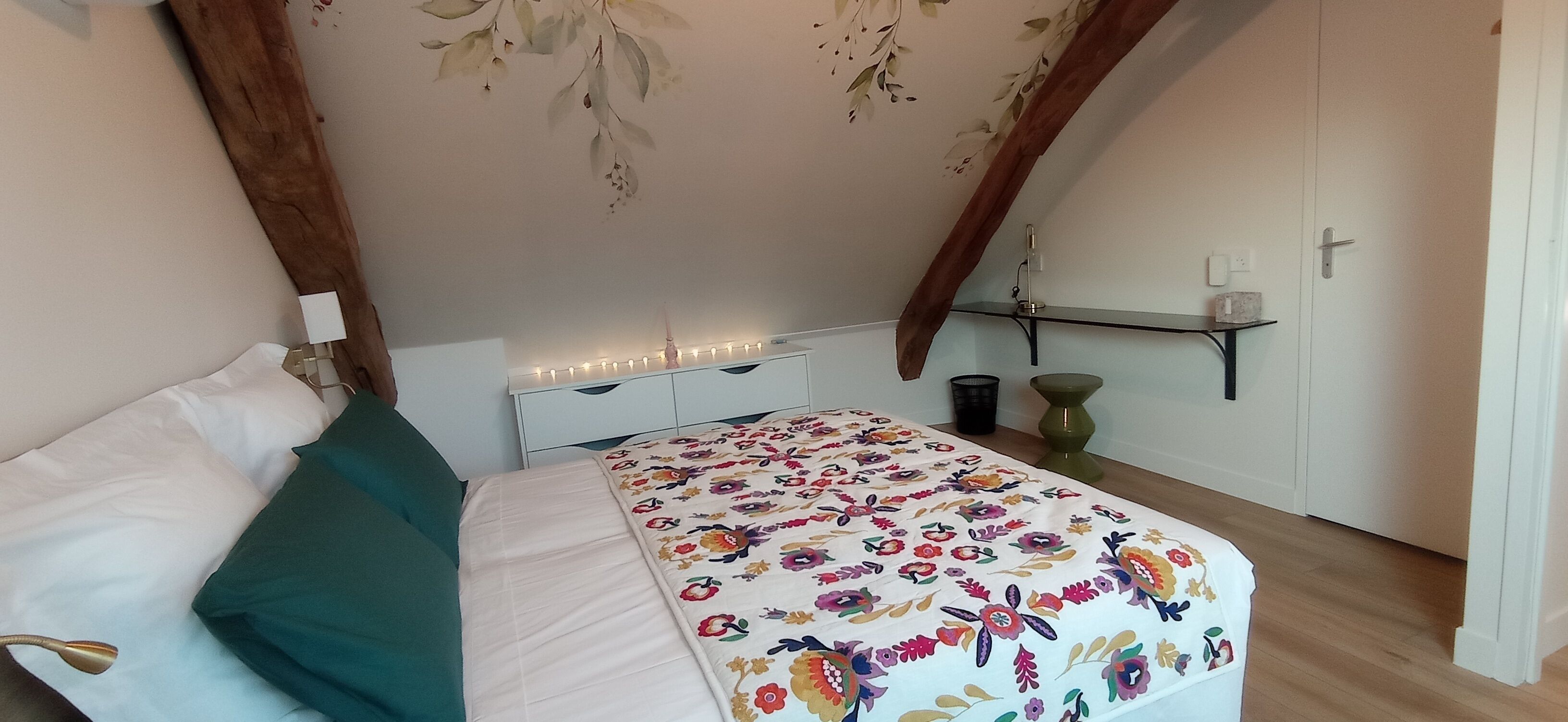 Quarto com cama, mesa de cabeceira, cômoda, prateleira na parede e porta.