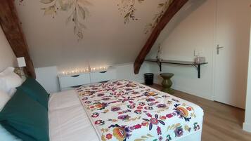 Quarto com cama, mesa de cabeceira, cômoda, prateleira na parede e porta.