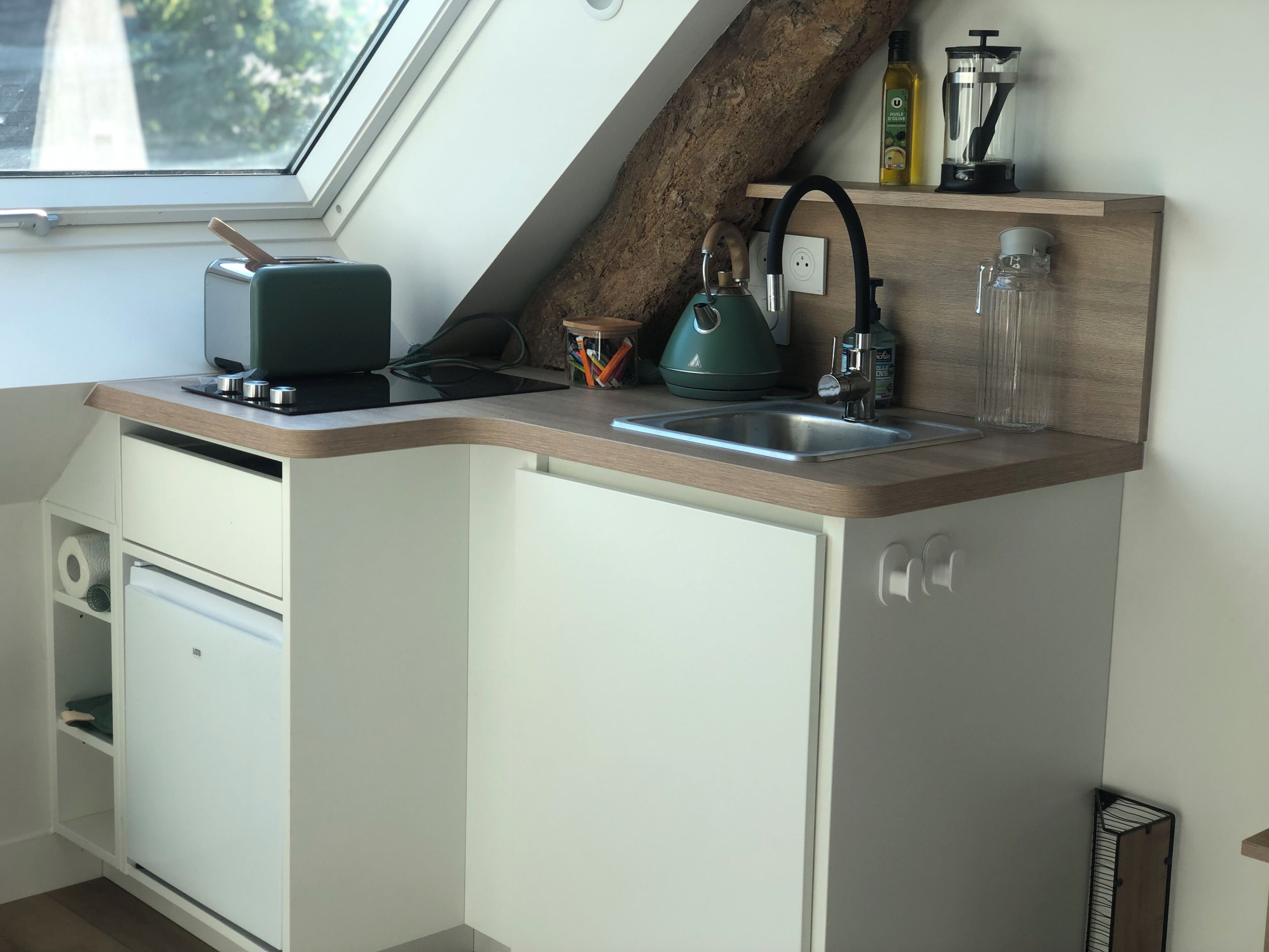 Een compacte keuken met een inbouw raam, een waterkoker, een koffiezetapparaat en een klein plankje met flessen.