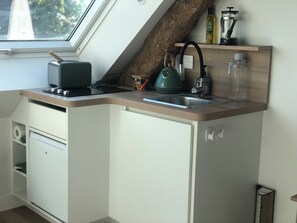 A compact kitchen with a built-in window, a kettle, a coffee maker, and a small shelf with bottles.