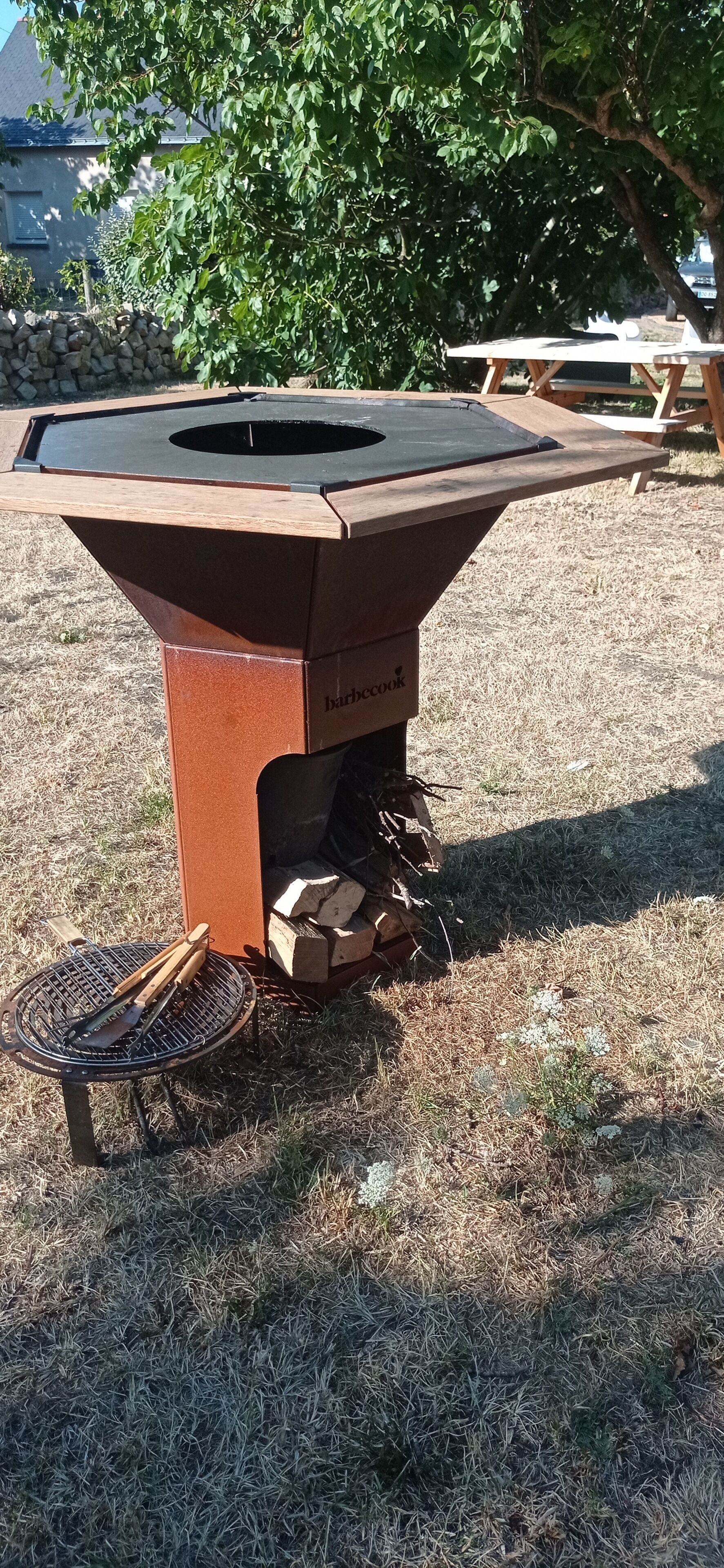 Un barbecue extérieur doté d’une table en métal et d’une cheminée, installé sur une pelouse, avec un mur de pierres et des arbres en arrière-plan.