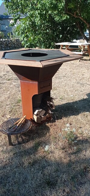 An outdoor barbecue grill with a metal table and a chimney, set on a grassy area with a stone wall and trees in the background. - Charming vacation rental with a jacuzzi/spa—Anjou (castles), countryside, river, hiking (Étriché)