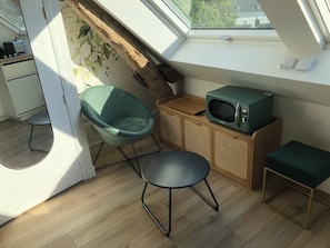 A compact living space with a skylight, a teal chair, a small table, a microwave, and a wooden cabinet.